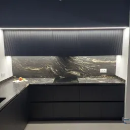 Czarna kuchnia z blatem z kamienia o unikalnym wzorze, podświetlane szafki górne z ryflowanym frontem, na blacie dekoracja w postaci owocu na białej desce.