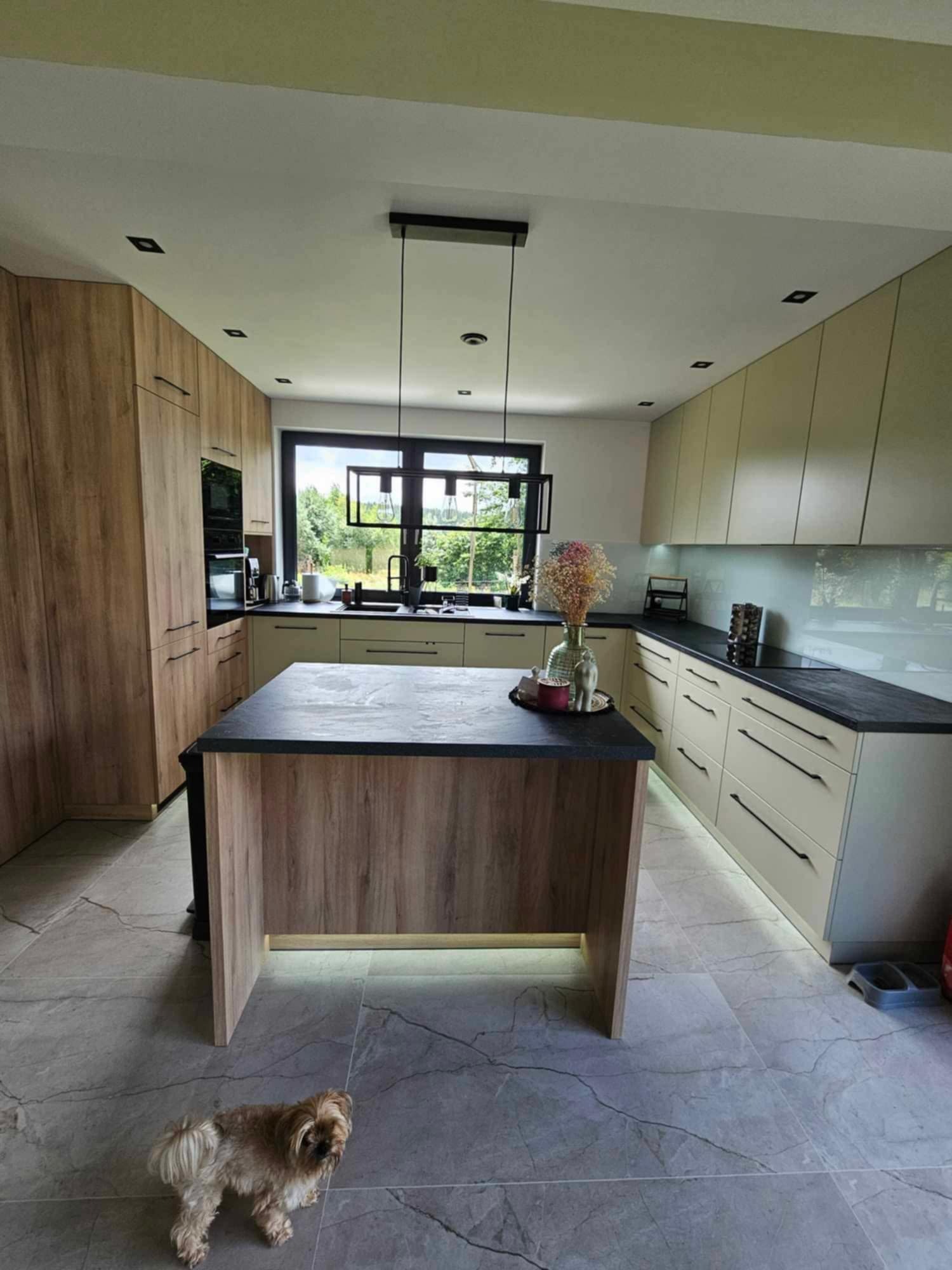 Nowoczesna kuchnia z wyspą, drewnianymi frontami i czarnymi blatami, oświetlenie LED pod wyspą, okno z widokiem na zieleń, mały pies na marmurowej podłodze.
