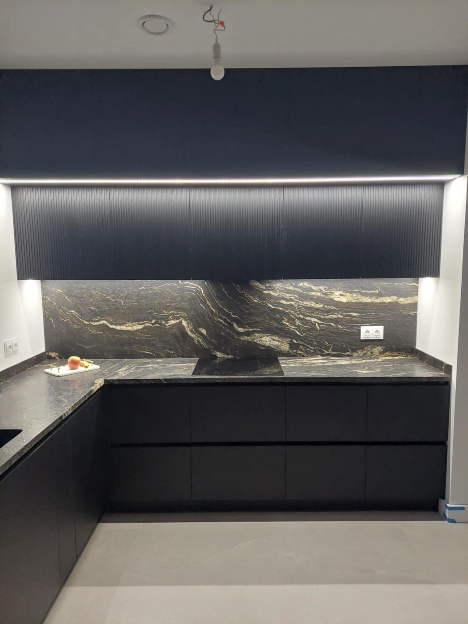 Czarna kuchnia z blatem z kamienia o unikalnym wzorze, podświetlane szafki górne z ryflowanym frontem, na blacie dekoracja w postaci owocu na białej desce.