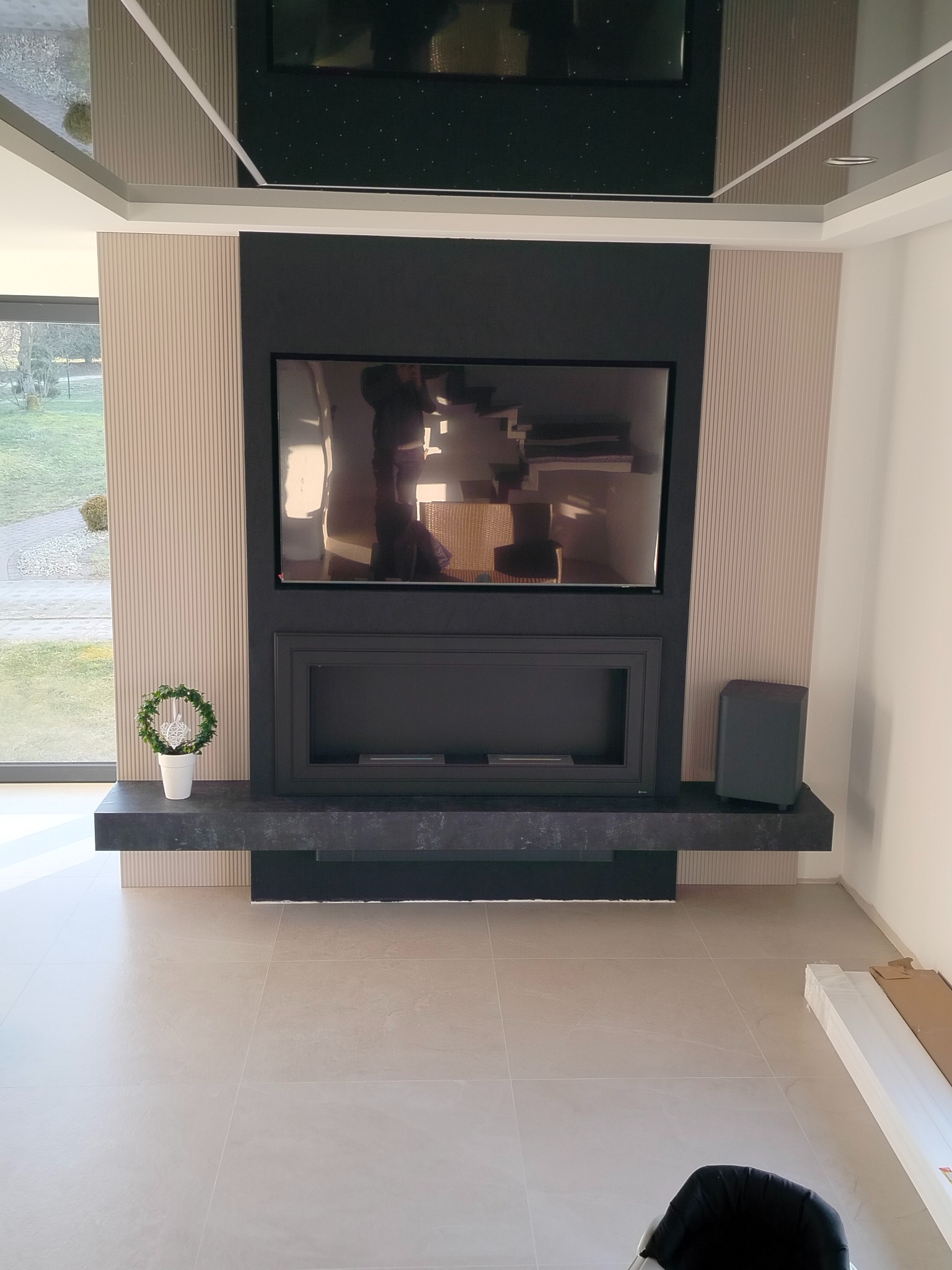 Nowoczesna aranżacja ściany z kominkiem i telewizorem, wykończona panelami dekoracyjnymi i blatem z ciemnego kamienia, uzupełniona minimalistyczną dekoracją roślinną.