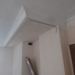 Maciej Szczęsny - Wnętrze w trakcie prac wykończeniowych: widoczne listwy sufitowe z zamontowanymi metalowymi profilami, ściany pokryte gładzią, karton z listwami przypodłogowymi oparty o ścianę.