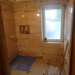 Maciej Szczęsny - Łazienka w trakcie układania płytek imitujących trawertyn, widoczne narzędzia glazurnicze, poziomica, worek z zaprawą i system poziomowania płytek z czerwonymi klipsami, mała wnęka w ścianie...