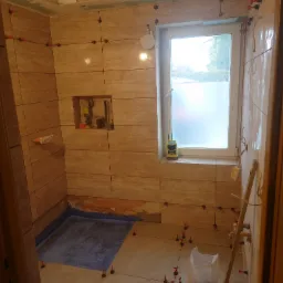 Łazienka w trakcie układania płytek imitujących trawertyn, widoczne narzędzia glazurnicze, poziomica, worek z zaprawą i system poziomowania płytek z czerwonymi klipsami, mała wnęka w ścianie...