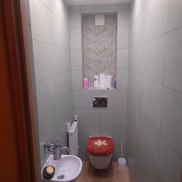 Maciej Szczęsny - Niewielka łazienka z umywalką na postumencie, toaletą z czerwoną nakładką i zielonymi płytkami, uzupełniona wnęką z tapetą w liście i drobnymi akcesoriami.