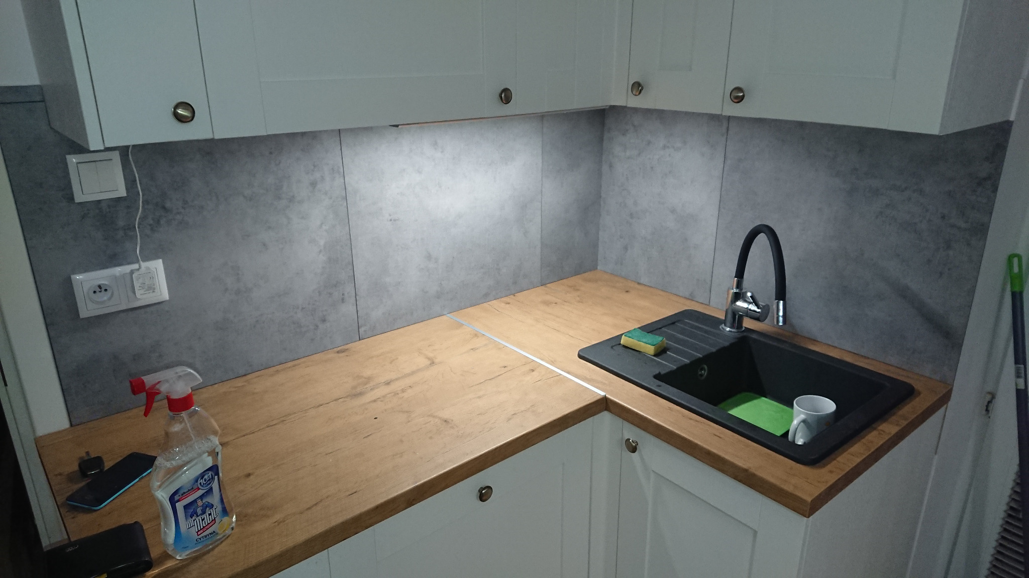 Nowoczesna kuchnia z drewnianym blatem, szarą ścianą i czarnym zlewozmywakiem; widoczny środek czyszczący i akcesoria.