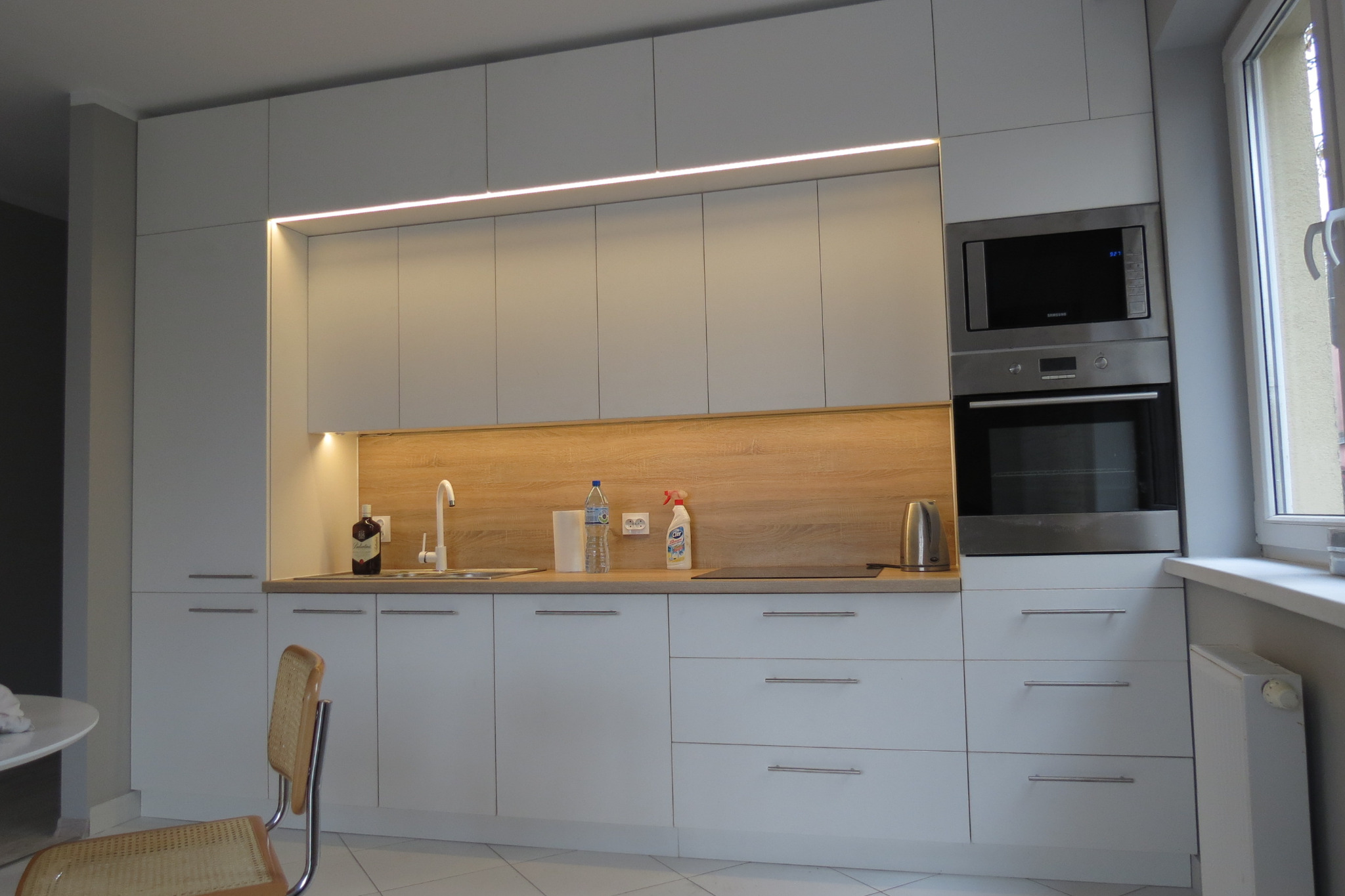 Nowoczesna kuchnia z białymi szafkami, drewnianym panelem nad blatem i oświetleniem LED podszafkowym, widoczny piekarnik i mikrofalówka w zabudowie oraz krzesło z plecionki.