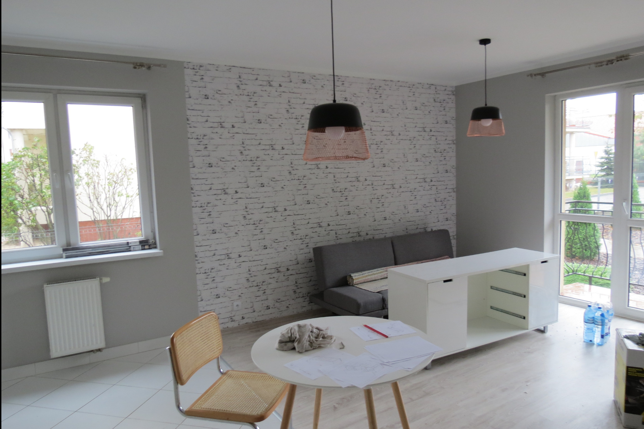 Pokój w trakcie wykańczania: szara sofa, biała komoda bez frontów, okrągły stół z planami, lampy wiszące z ażurowymi abażurami, ściana z imitacją białej cegły.