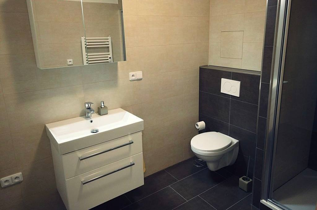 Wykończona łazienka we Wrocławiu z beżowymi płytkami na ścianach, ciemnymi płytkami podłogowymi, białą umywalką z szafką, wiszącą toaletą i kabiną prysznicową z metalowymi ramami.