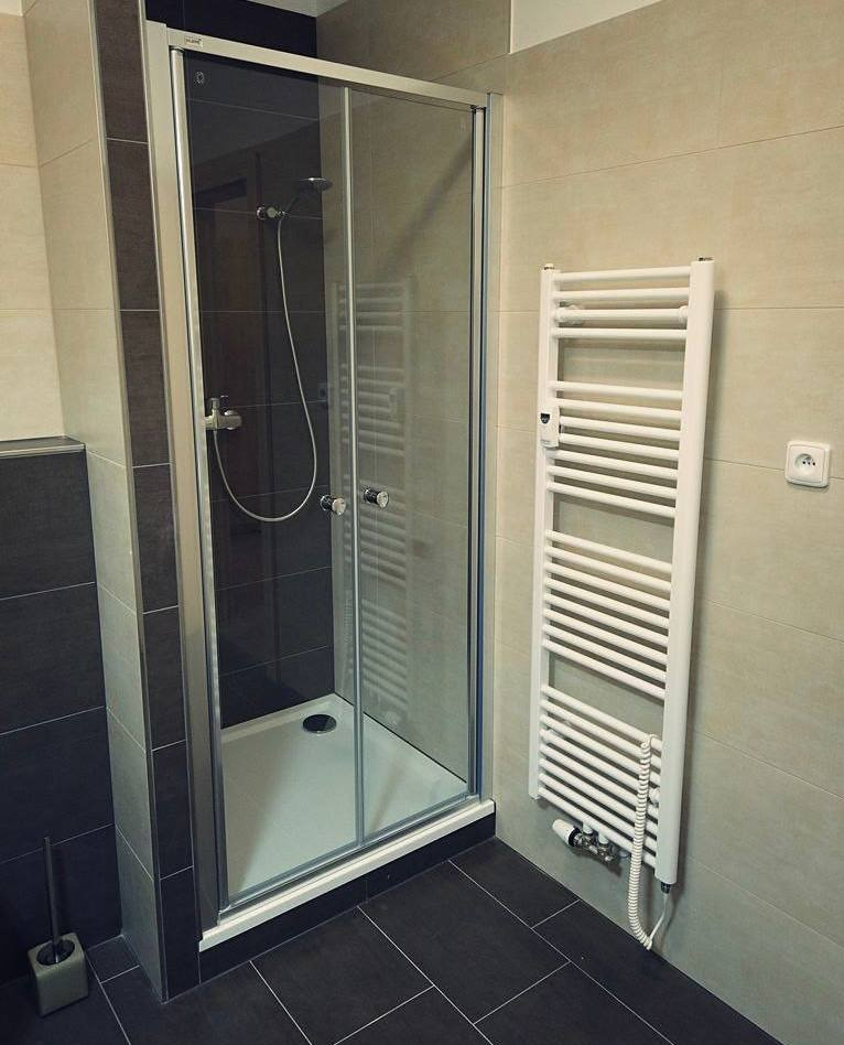 Nowoczesna kabina prysznicowa z przesuwnymi drzwiami, ciemnoszare płytki podłogowe i ścienne, biały grzejnik drabinkowy na beżowej ścianie.