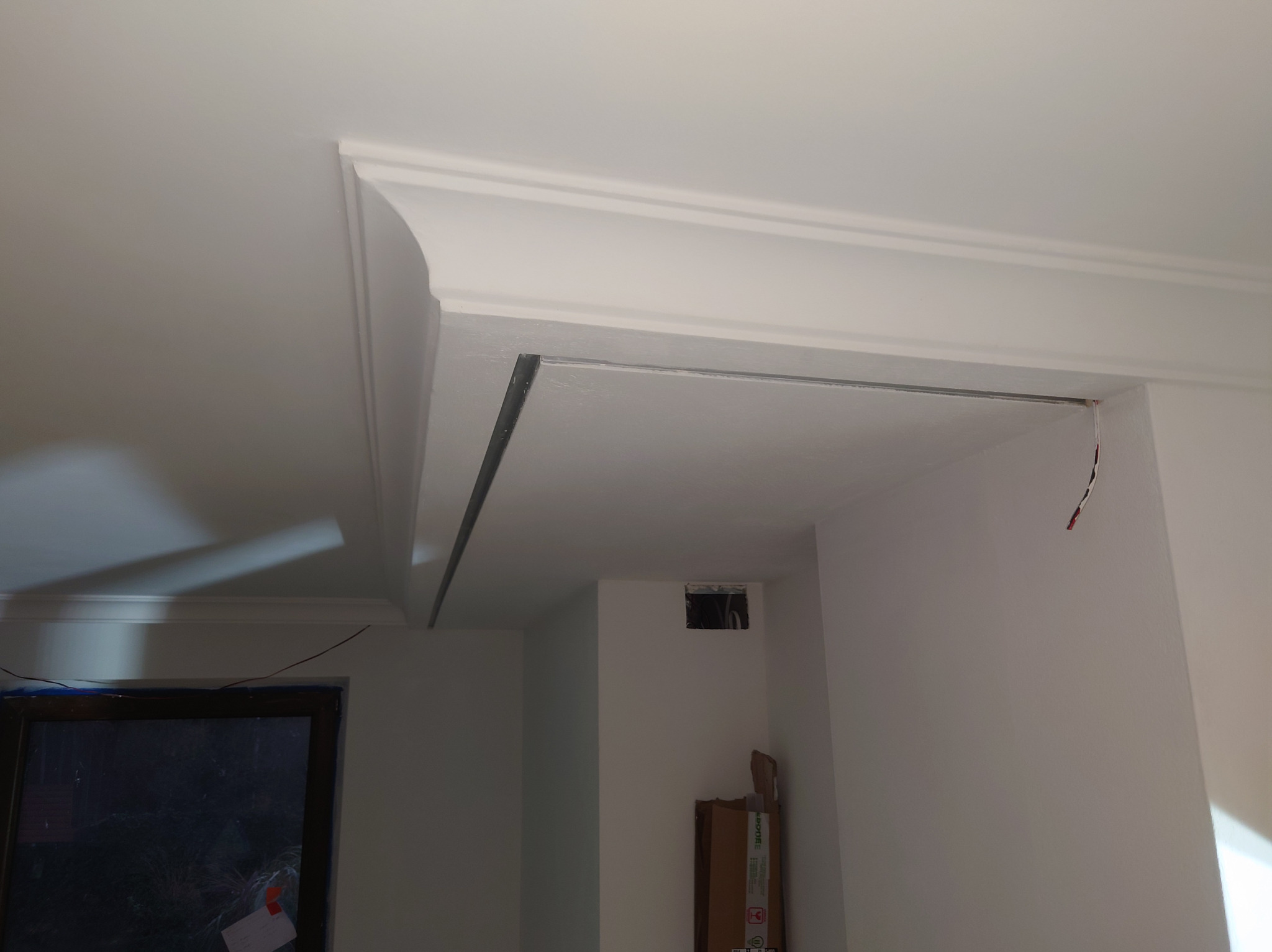 Wnętrze w trakcie prac wykończeniowych: widoczny fragment sufitu z zamontowaną listwą oświetleniową i dekoracyjnym gzymsem, wystające przewody elektryczne, okno zabezpieczone folią malarską.