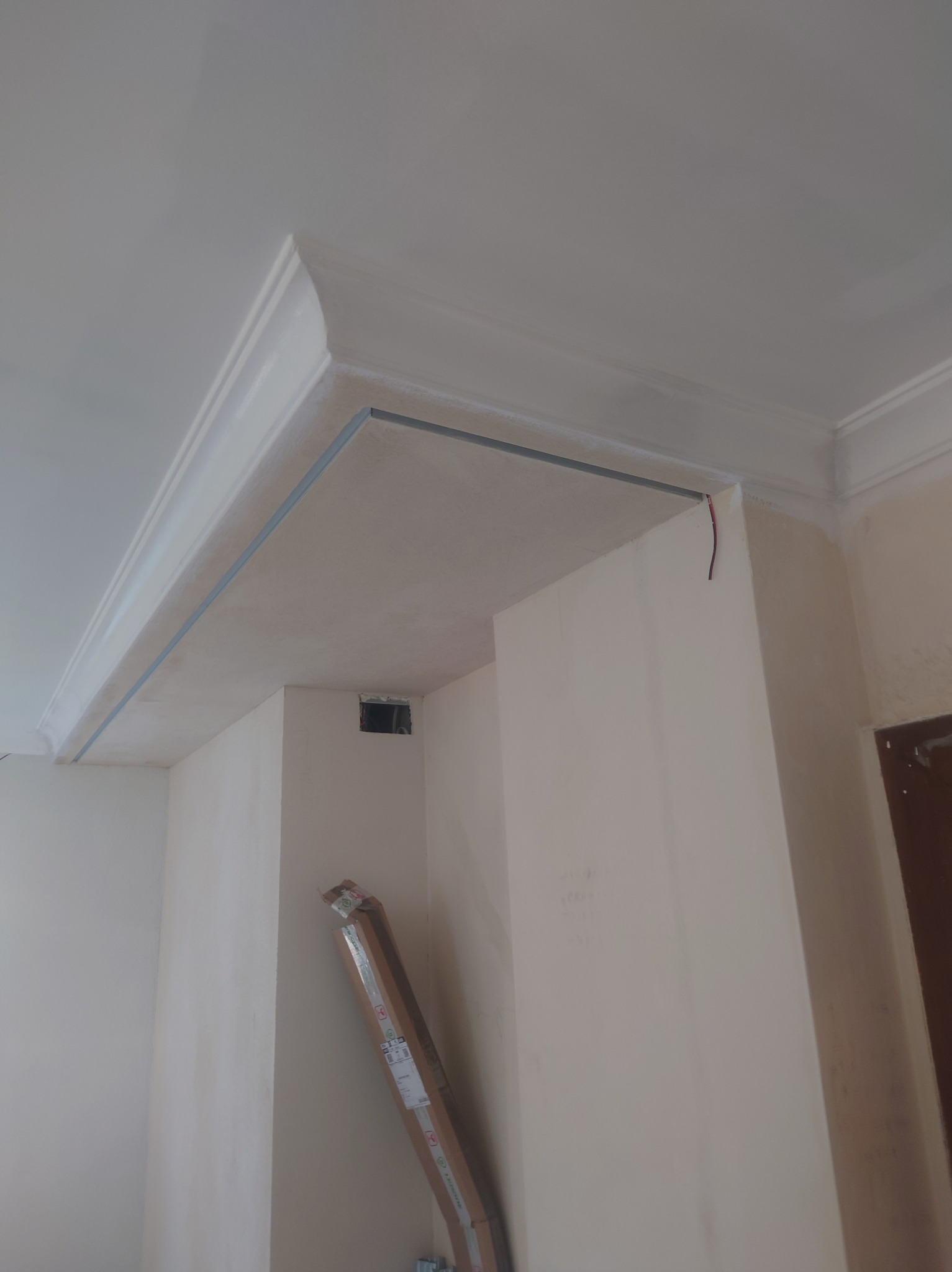 Wnętrze w trakcie prac wykończeniowych: widoczne listwy sufitowe z zamontowanymi metalowymi profilami, ściany pokryte gładzią, karton z listwami przypodłogowymi oparty o ścianę.