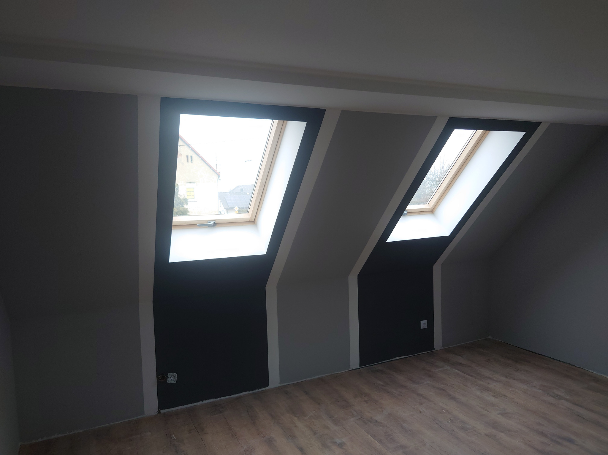 Poddasze z dwoma oknami dachowymi po malowaniu ścian na szaro i czarno z panelami podłogowymi o wzorze drewna.
