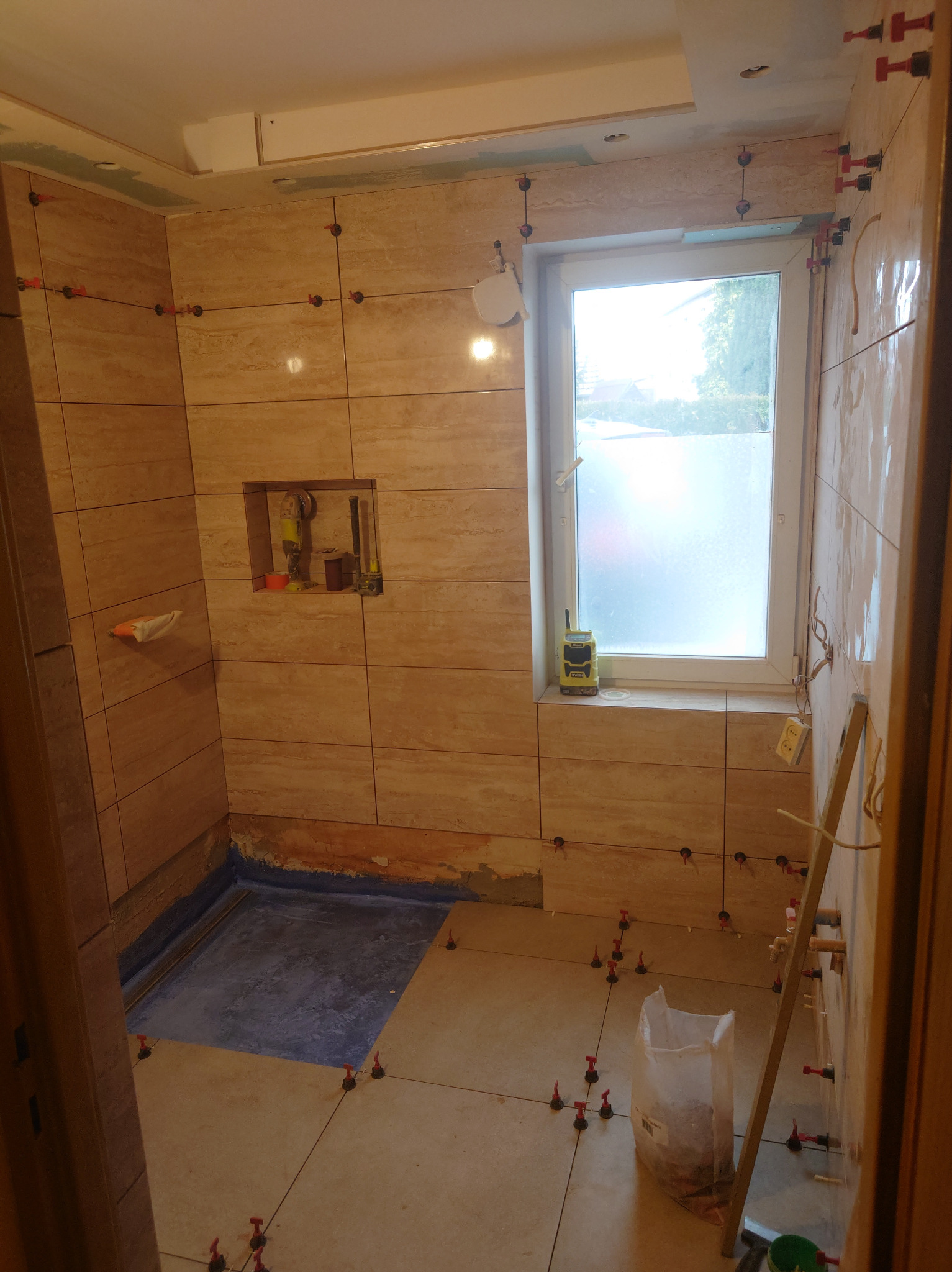 Łazienka w trakcie układania płytek imitujących trawertyn, widoczne narzędzia glazurnicze, poziomica, worek z zaprawą i system poziomowania płytek z czerwonymi klipsami, mała wnęka w ścianie...