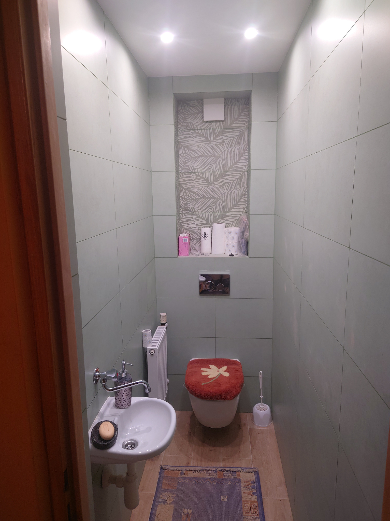 Niewielka łazienka z umywalką na postumencie, toaletą z czerwoną nakładką i zielonymi płytkami, uzupełniona wnęką z tapetą w liście i drobnymi akcesoriami.