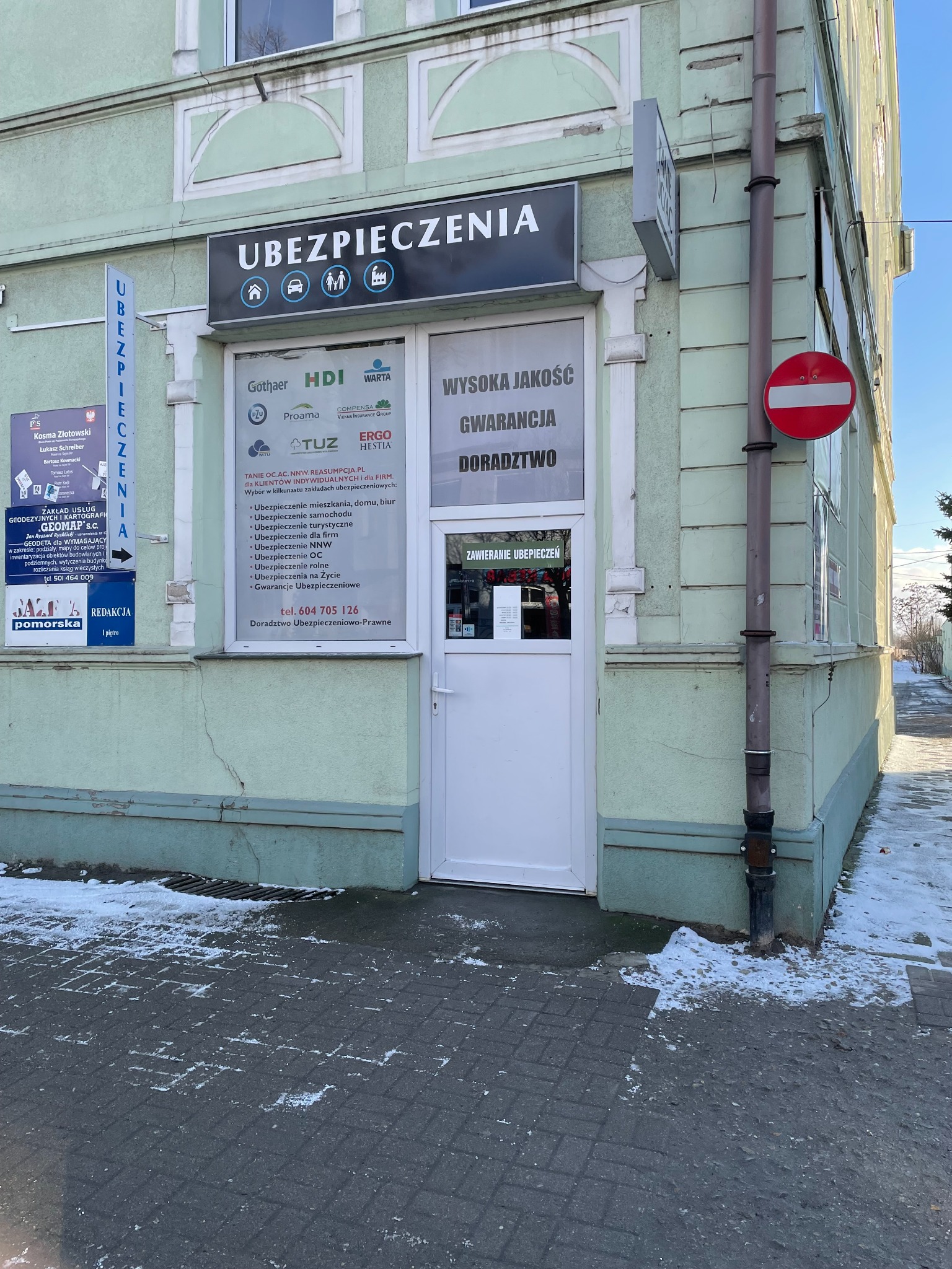 Fasada budynku z szyldem 'Ubezpieczenia' i logo różnych firm ubezpieczeniowych na witrynie, znak zakazu wjazdu na rogu.
