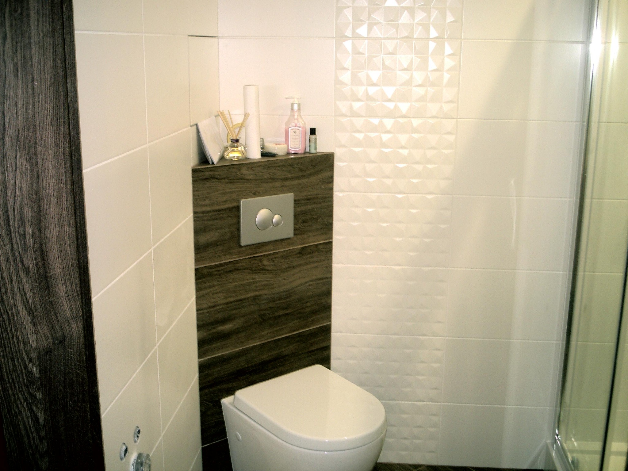 Nowoczesna toaleta podwieszana z szarym panelem drewnopodobnym i przyciskiem spłukującym, otoczona białymi płytkami z trójwymiarowym wzorem oraz elementami dekoracyjnymi na półce.