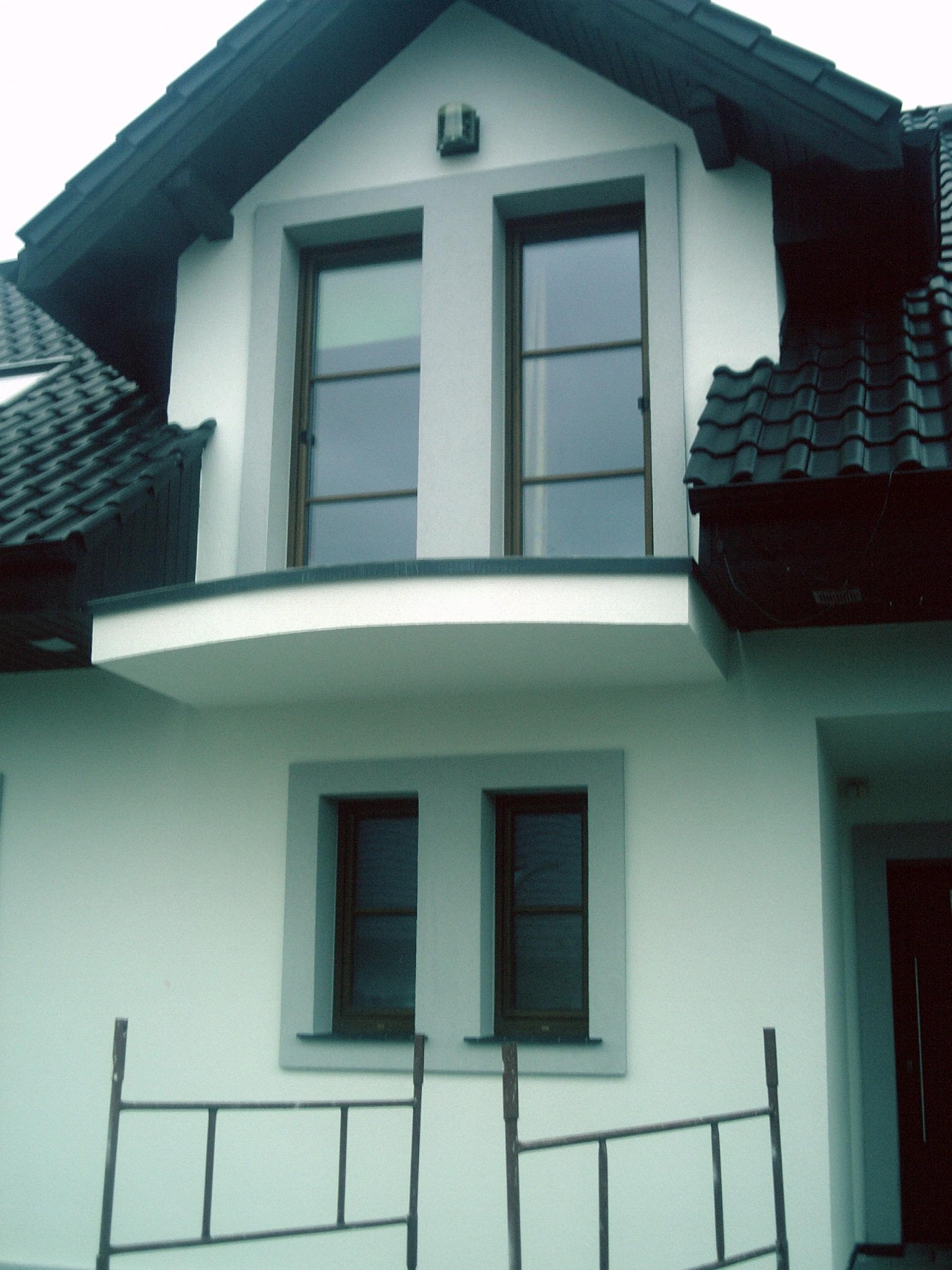 Elewacja domu z dwoma oknami na górze i dwoma na dole, jasna ściana, ciemna dachówka, widoczne rusztowanie w dolnej części kadru.