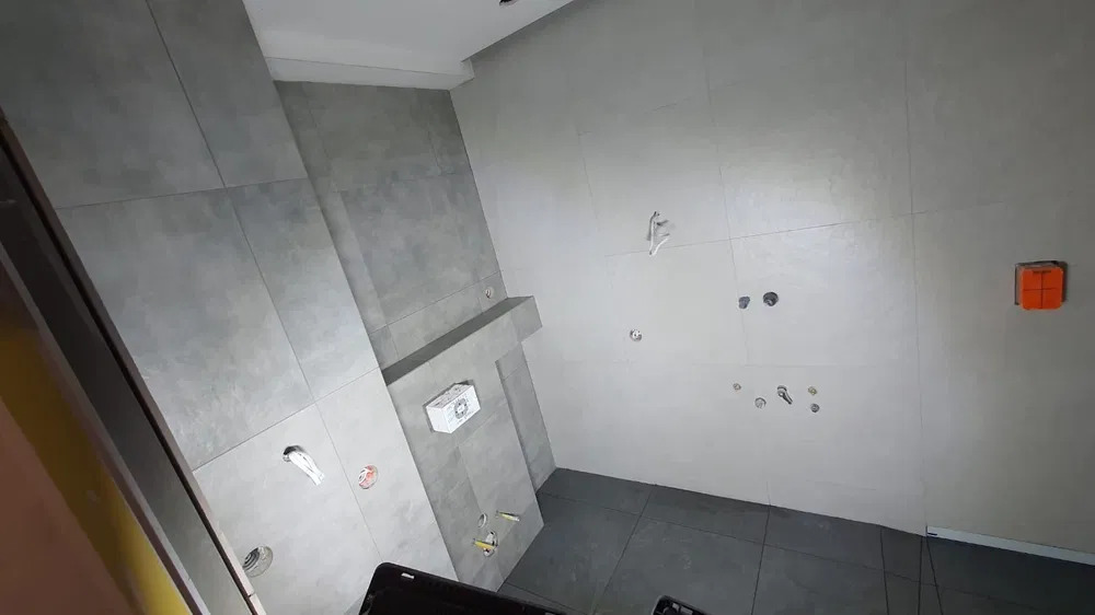 Szare płytki wielkoformatowe na ścianach i podłodze w surowej łazience, widoczne otwory na instalacje, wystające kable i puszka elektryczna