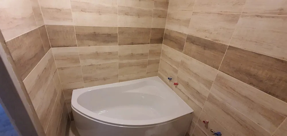 Narożna wanna w obudowie z płytek imitujących drewno, widoczne zawory ciepłej i zimnej wody.