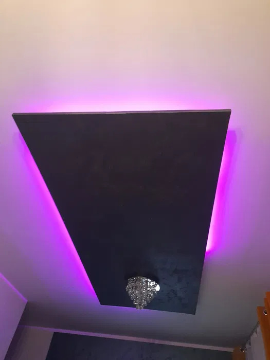 Ciemna, trapezoidalna płyta sufitowa z podświetleniem LED w kolorze fioletowym i małym, kryształowym żyrandolem pośrodku.