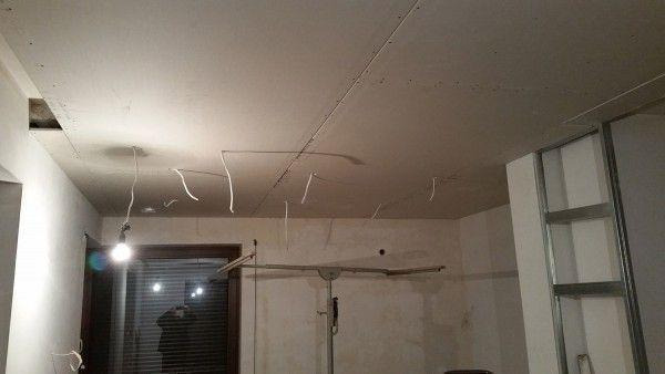 Pokój w trakcie remontu: sufit z płyt gipsowo-kartonowych z widocznymi przewodami, metalowa konstrukcja ścianki działowej, lampa wisząca na kablu i statyw z poprzeczką.