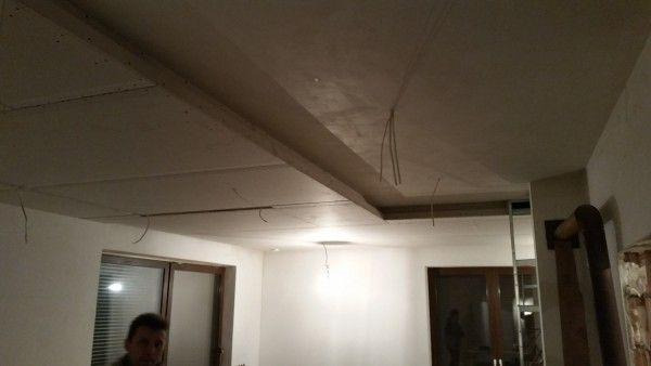 Niedokończony sufit z płyt gipsowo-kartonowych z wystającymi przewodami, widoczne elementy konstrukcyjne i ślady niedawnej pracy, fragment twarzy osoby w lewym dolnym rogu.