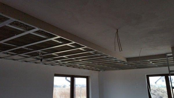 Metalowa konstrukcja sufitu podwieszanego w trakcie montażu, widoczne okna z widokiem na zewnątrz, surowy stan wykończenia wnętrza.