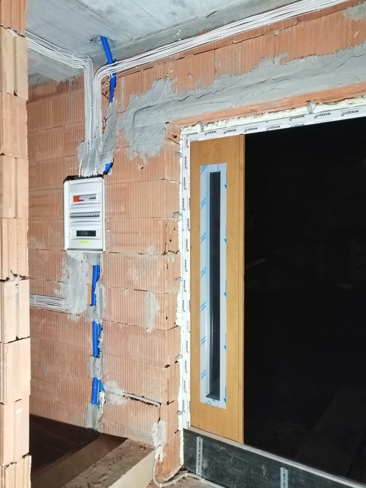 Niedokończona instalacja elektryczna i okno w surowym stanie, widoczne przewody w peszlach, skrzynka elektryczna na ścianie z cegły dziurawki, okno z folią ochronną na ramie.