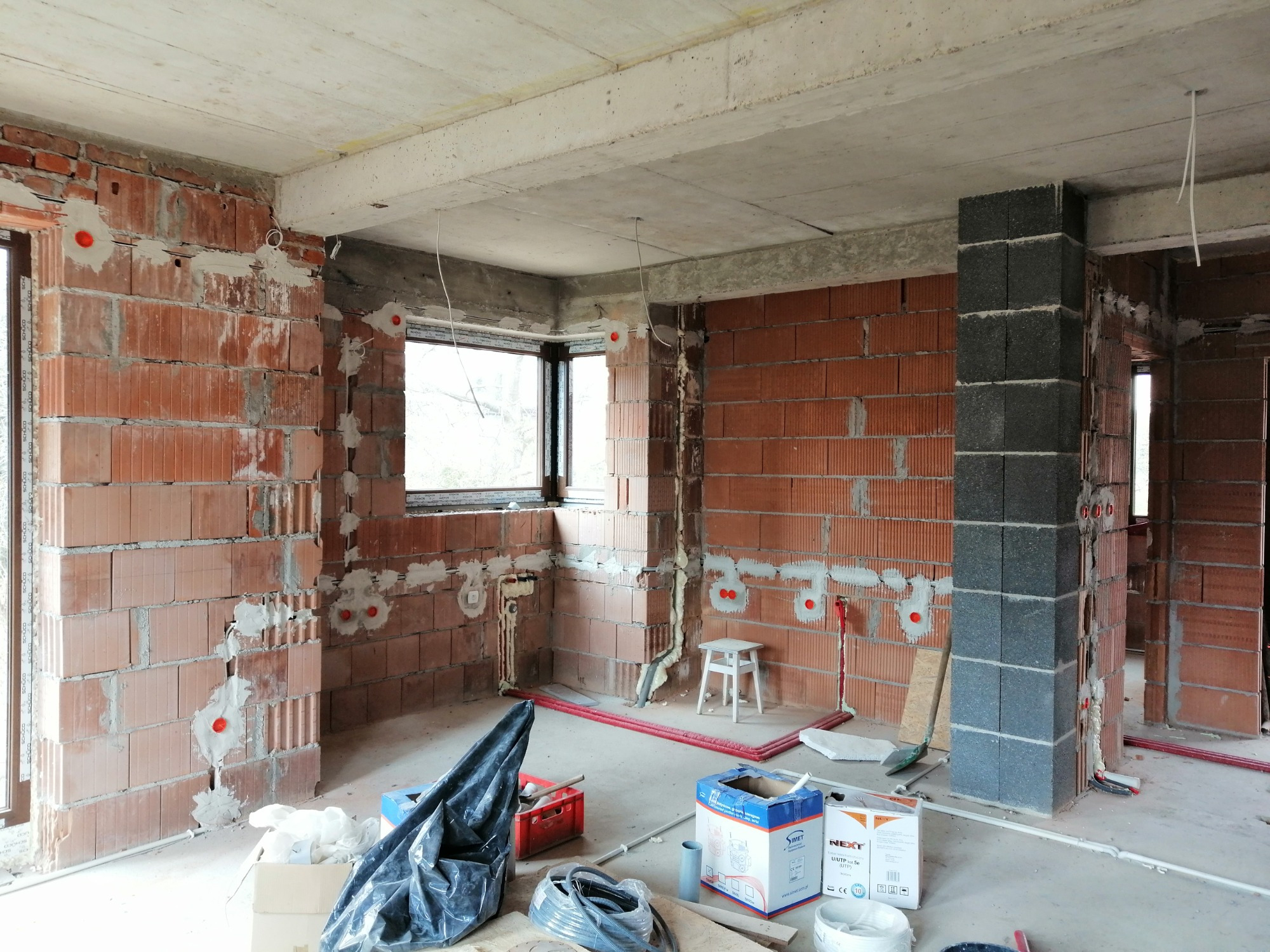 Surowy stan wnętrza budynku w trakcie prac instalacyjnych: widoczne ceglane ściany z wkuciami pod instalacje elektryczne i hydrauliczne, betonowy strop, okno, częściowo wykończony filar z szarych...
