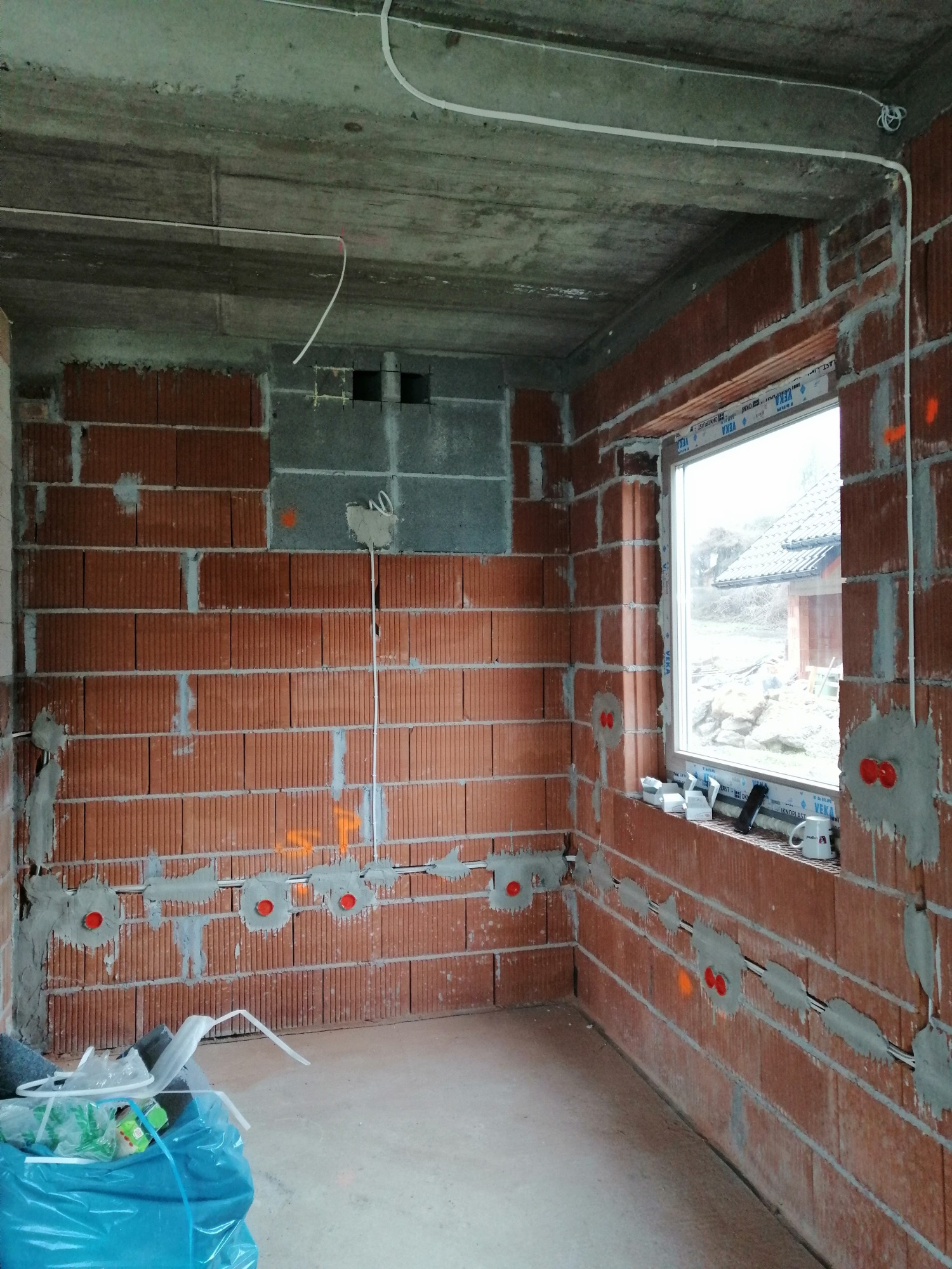 Surowy stan wnętrza w trakcie prac instalacyjnych: ceglane ściany z widocznymi przewodami elektrycznymi w szarym tynku, surowy betonowy strop, okno z widokiem na zewnątrz, śmieci w worku na podłodze.