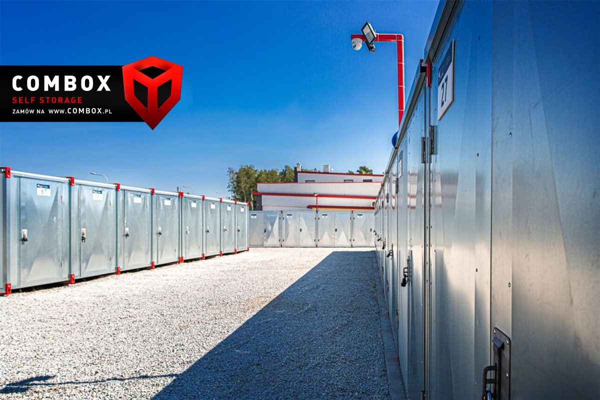 Rząd metalowych kontenerów samoobsługowych z numerem 27, oświetlonych kamerą przemysłową na tle błękitnego nieba i żwirowego placu.