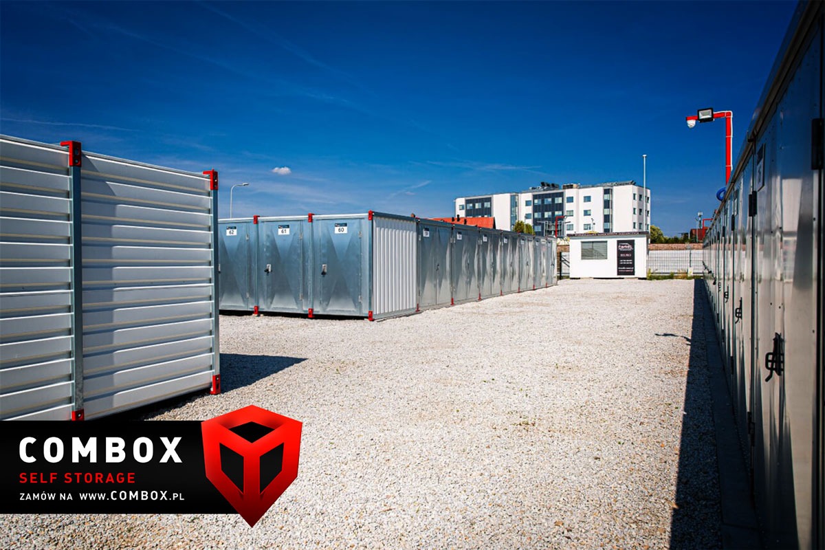 Rząd ponumerowanych, metalowych kontenerów do samodzielnego składowania na żwirowym placu, z budynkiem w tle i logo firmy w rogu.