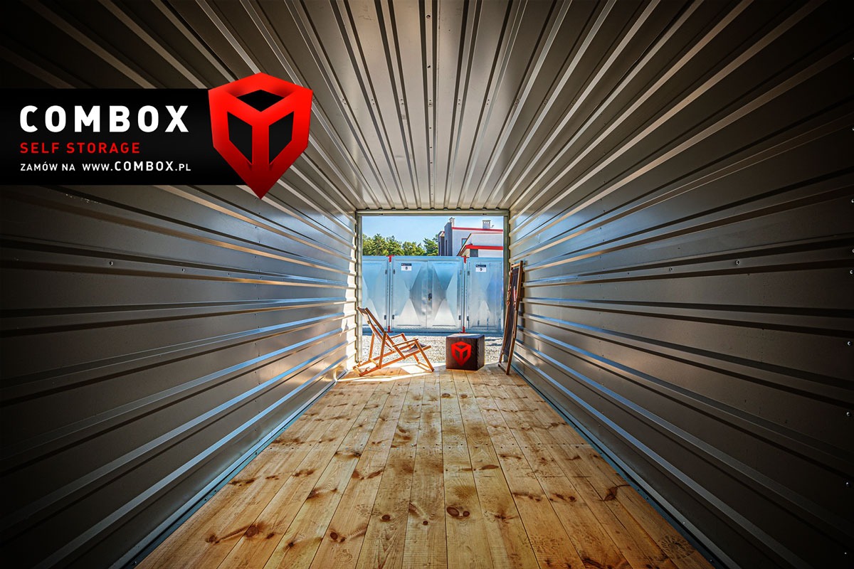 Wnętrze kontenera z drewnianą podłogą, widok na zewnątrz przez otwarte drzwi, krzesło plażowe i kostka z logo.