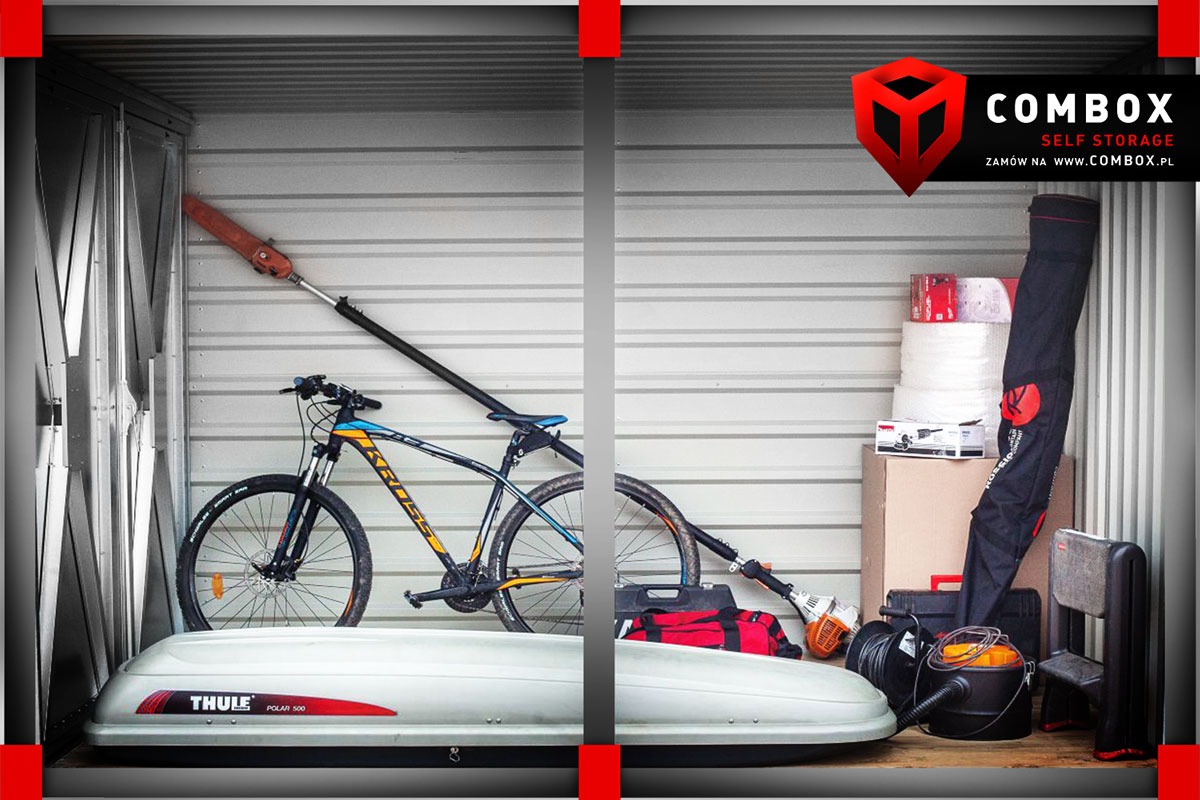 Wnętrze boksu magazynowego z rowerem Kross, białym boxem dachowym Thule Polar 500, narzędziami ogrodniczymi i innymi przedmiotami, widoczny logotyp Combobox Self Storage w prawym górnym rogu.