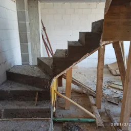 Surowe betonowe schody w trakcie budowy, widoczne drewniane szalunki i podpory, ściany z bloczków betonu komórkowego w tle, na podłodze gruz i resztki materiałów budowlanych.