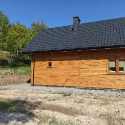 Nowoczesny dom szkieletowy z drewnianą elewacją, ciemnym dachem z blachodachówki i czarnym kominem, otoczony zielenią i przygotowanym terenem pod ogród.