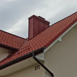 Czerwona dachówka falista na skośnym dachu domu z beżową elewacją i ciemnym kominem, widoczne rynny i numer domu 271.