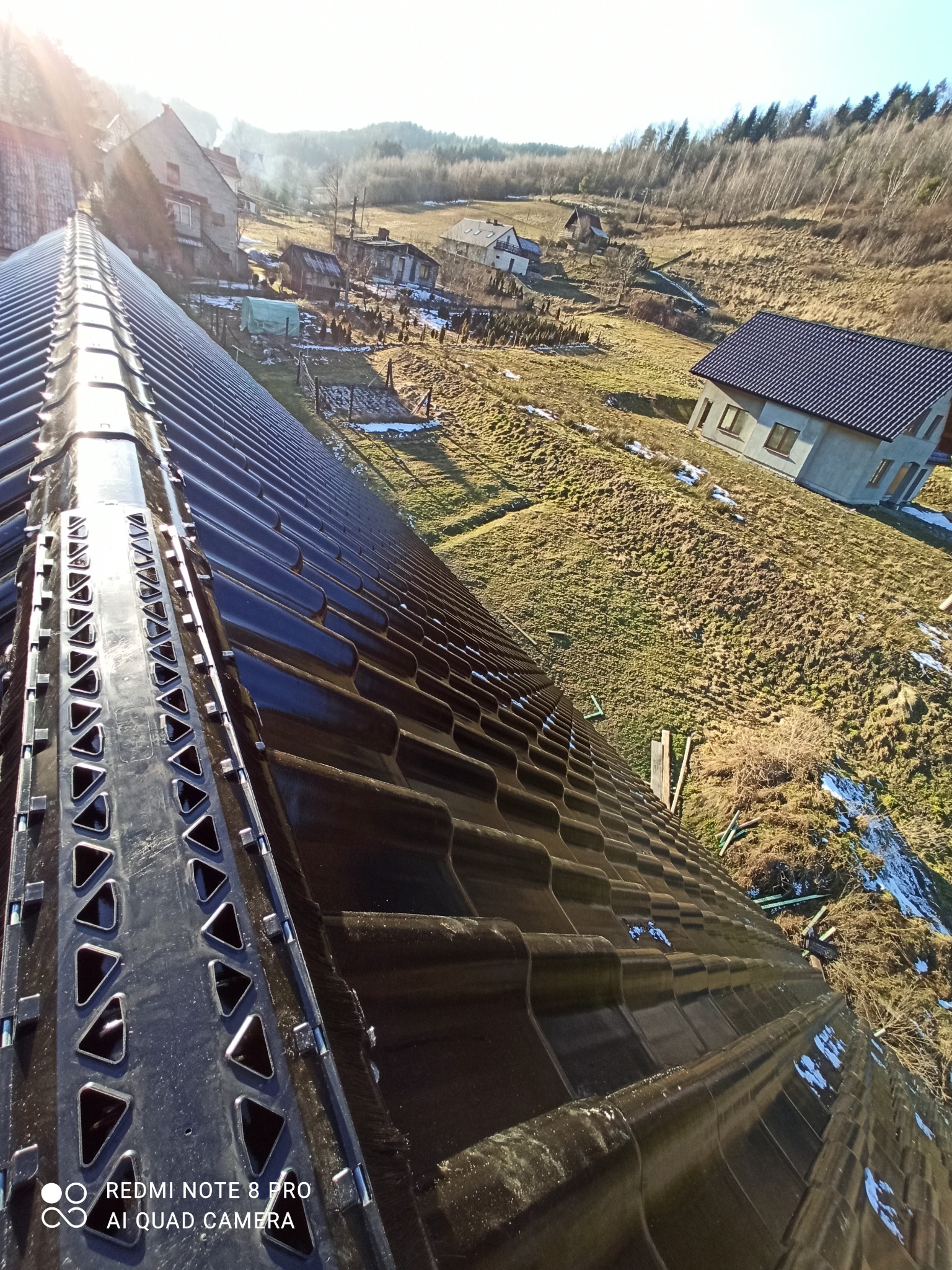 Nowe, błyszczące, czarne dachówki ceramiczne z elementami wentylacyjnymi, widok z góry na tle wiejskiego krajobrazu z domami i pagórkami w słoneczny dzień.