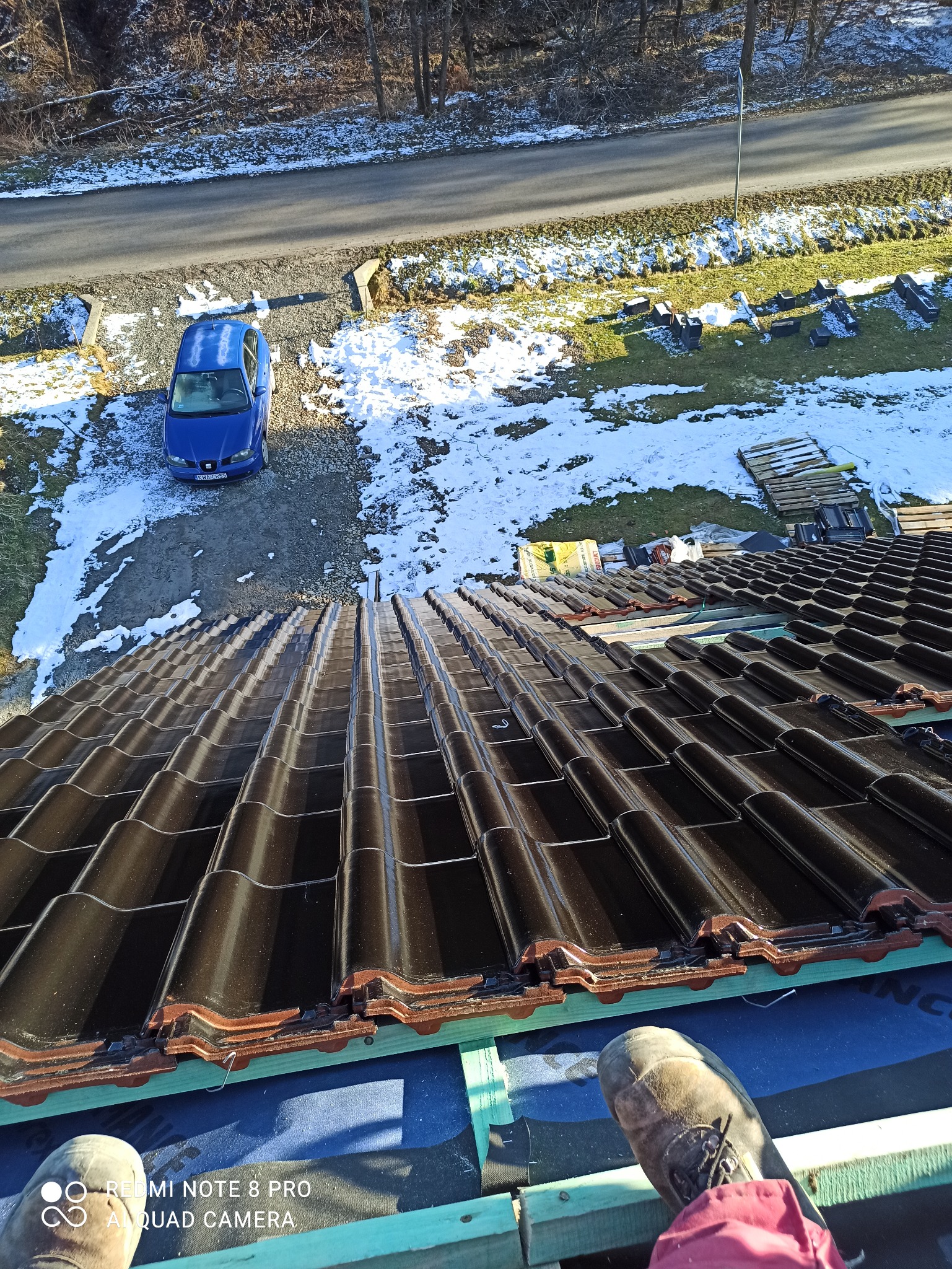 Widok z góry na nowo ułożone, błyszczące, ciemnobrązowe dachówki ceramiczne na skośnym dachu w trakcie montażu, widoczne fragmenty łat i kontrłat, w tle błękitny samochód osobowy i resztki śniegu...
