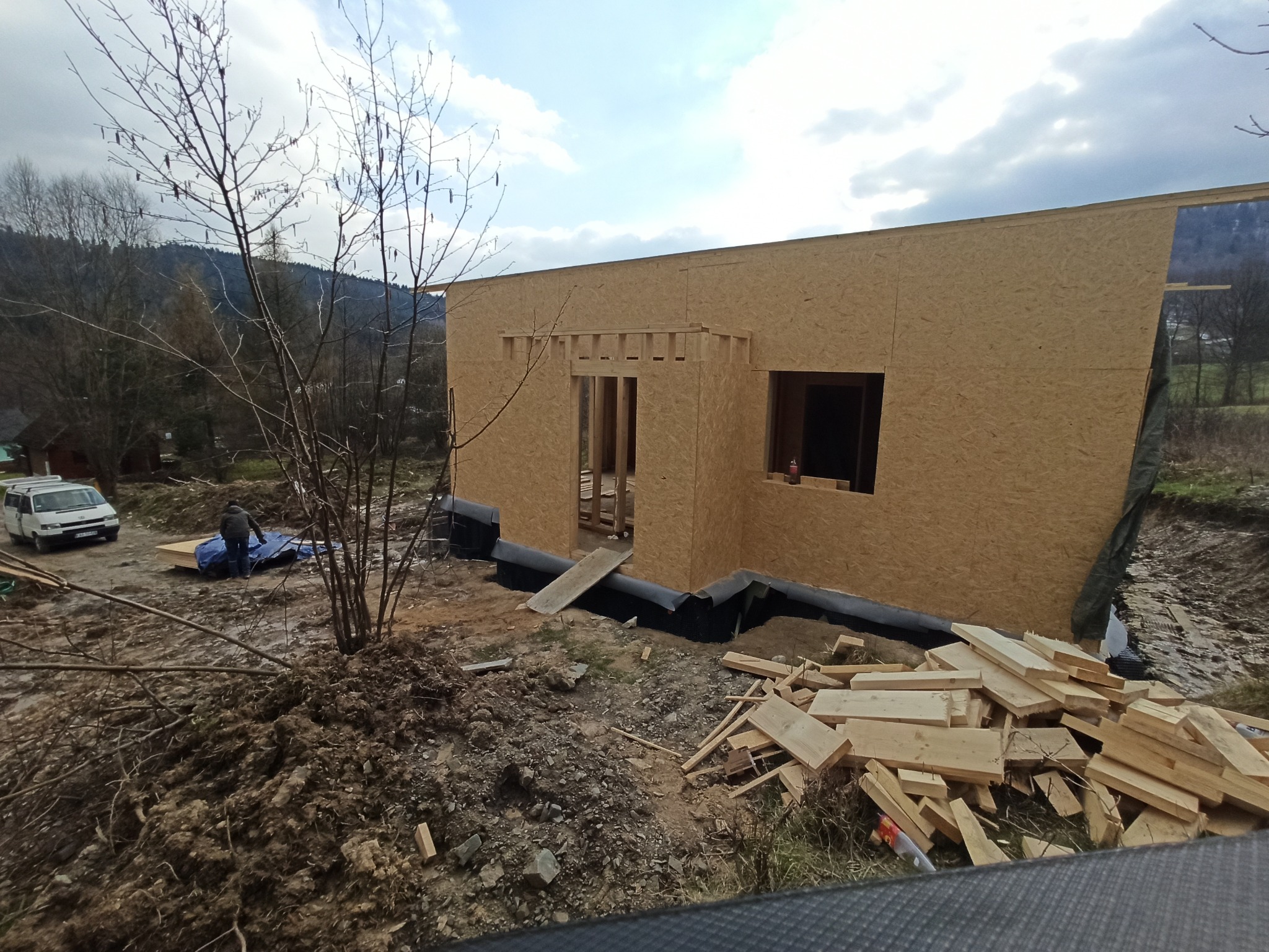 Wznoszenie domu szkieletowego z płyt OSB, widoczne drewniane elementy konstrukcyjne, izolacja fundamentów, teren budowy z ziemią i stertą drewna, w tle zalesione wzgórza i pochmurne niebo.