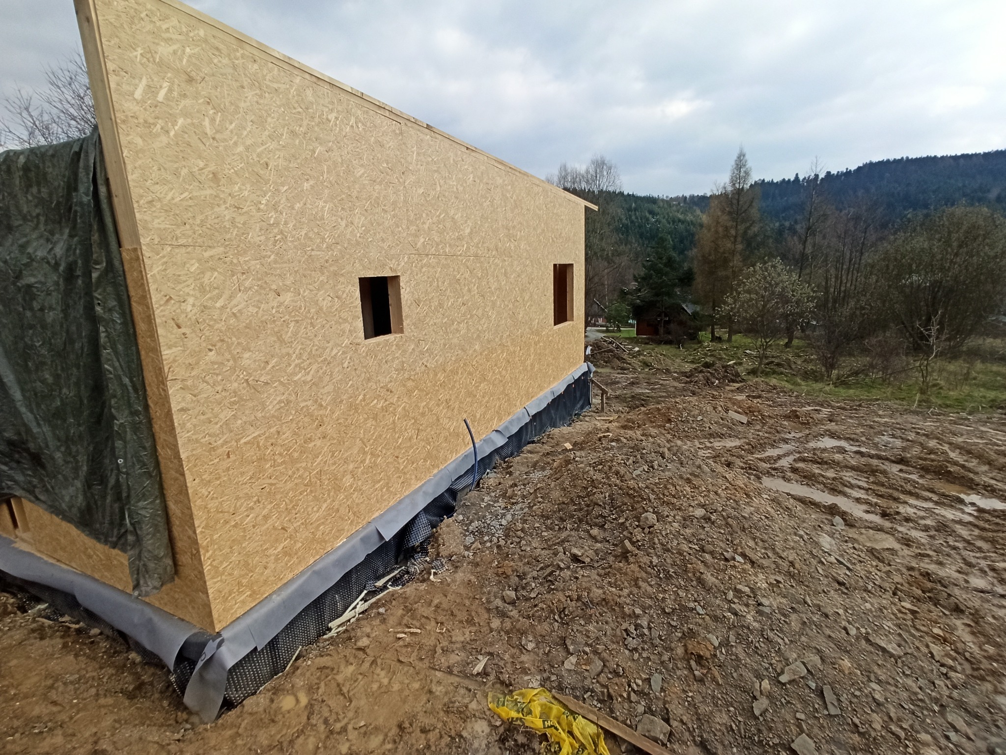 Wznoszenie ścian zewnętrznych domu szkieletowego z płyt OSB, widoczne otwory okienne, zabezpieczenie fundamentów czarną folią, w tle zalesione wzgórza.