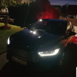 Czarny Ford Mondeo z włączonymi światłami przednimi, zaparkowany na tle domu i krzewów podświetlonych czerwoną lampą, widoczne tablice rejestracyjne.