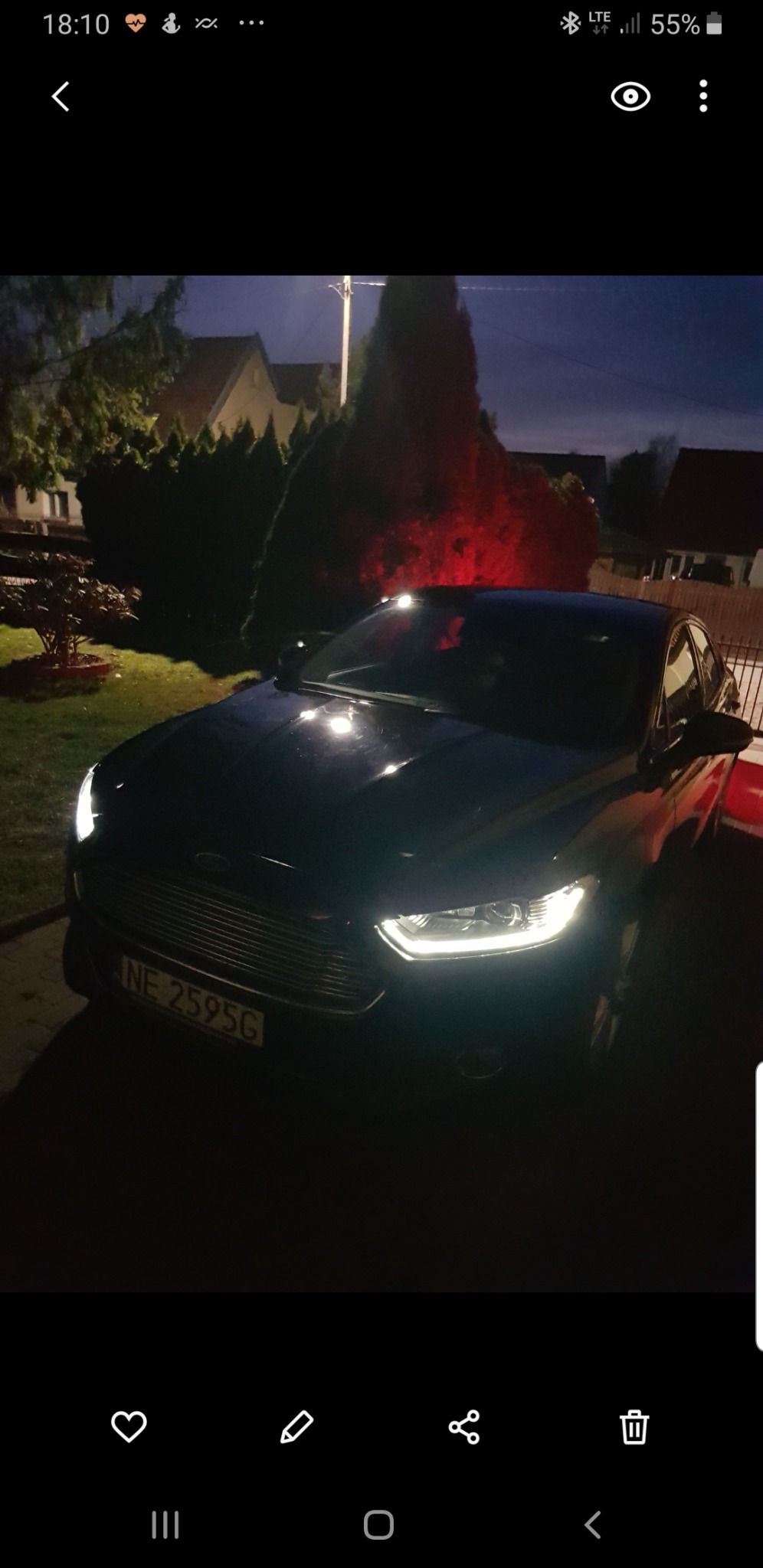 Czarny samochód osobowy Ford z włączonymi światłami mijania, zaparkowany na trawniku przed domem po zmroku.