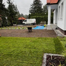 Układanie trawy z rolki na przygotowanym gruncie przed domem, widoczny wózek z trawą, auto firmy ogrodniczej w tle, częściowo ułożona trawa z widocznym systemem siatki wzmacniającej.