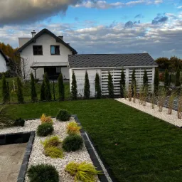 Nowoczesny ogród z równo przystrzyżonym trawnikiem, ozdobnymi rabatami wysypanymi białym kamieniem, niskimi krzewami i strzyżonymi tujami wzdłuż ogrodzenia oraz budynku gospodarczego.