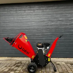 Czerwona, spalinowa rębarka do gałęzi marki Cedrus, stojąca na kostce brukowej przed szarą bramą garażową z widoczną lampą nad nią.