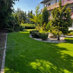 Dom i Ogród - Starannie utrzymany trawnik z równo przystrzyżoną trawą, obok ozdobne drzewa i krzewy oraz jasne kamienie dekoracyjne, na tle budynku z balkonami w słoneczny dzień.