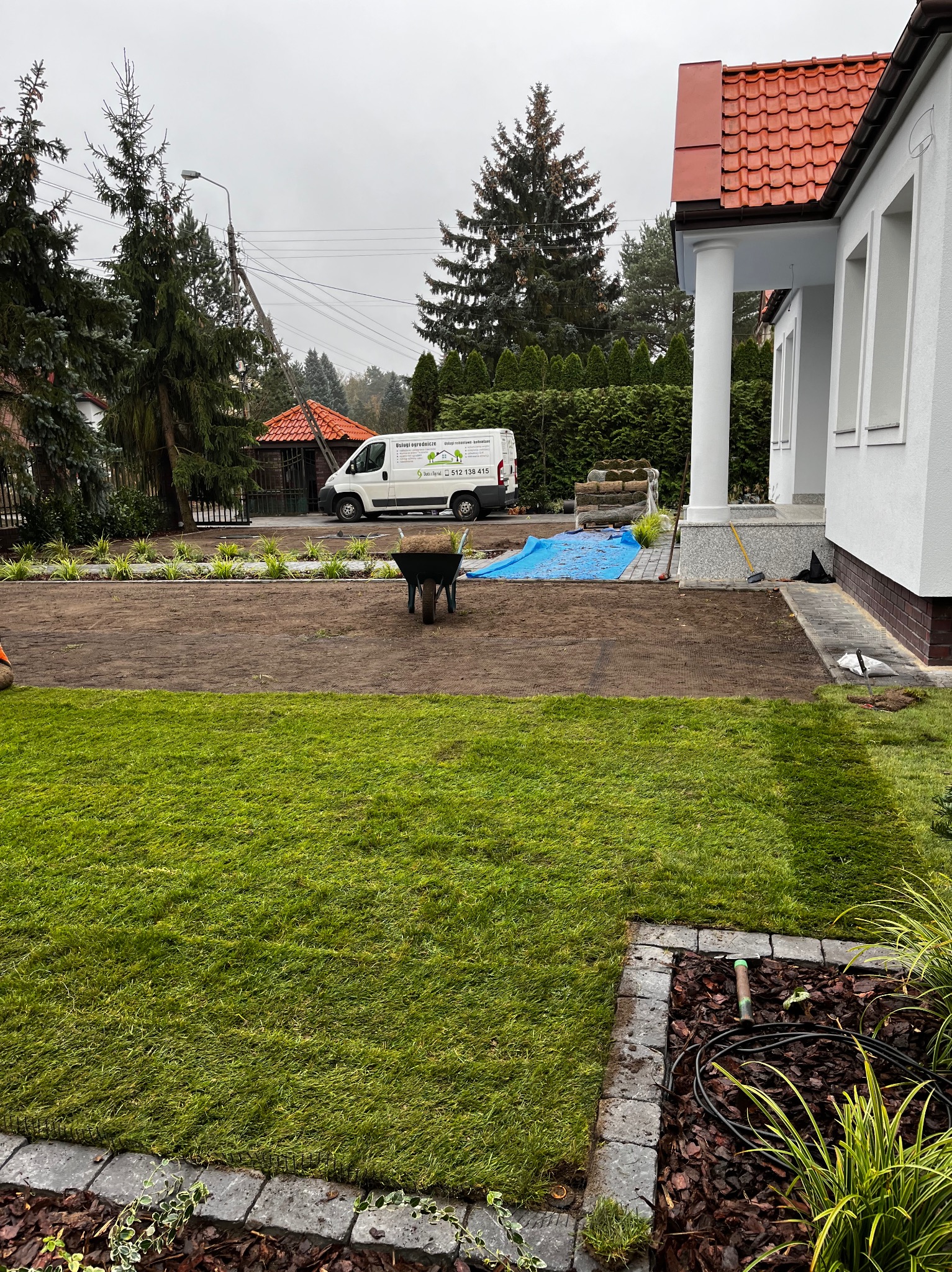Układanie trawy z rolki na przygotowanym gruncie przed domem, widoczny wózek z trawą, auto firmy ogrodniczej w tle, częściowo ułożona trawa z widocznym systemem siatki wzmacniającej.