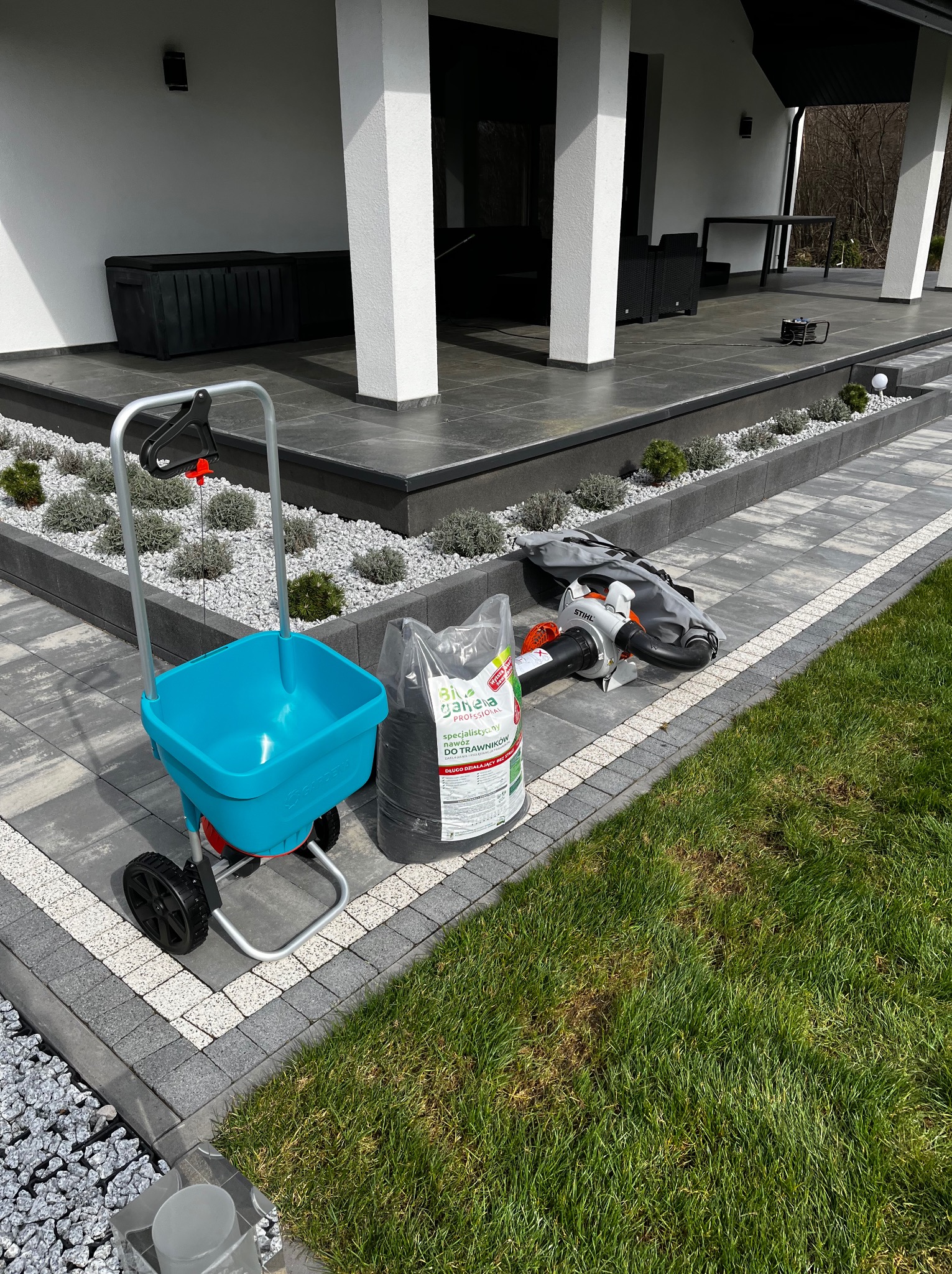 Niebieski rozsiewacz nawozu Gardena, worek nawozu Biopon i dmuchawa Stihl na kostce brukowej obok trawnika z widocznymi uszkodzeniami, na tle nowoczesnego domu.