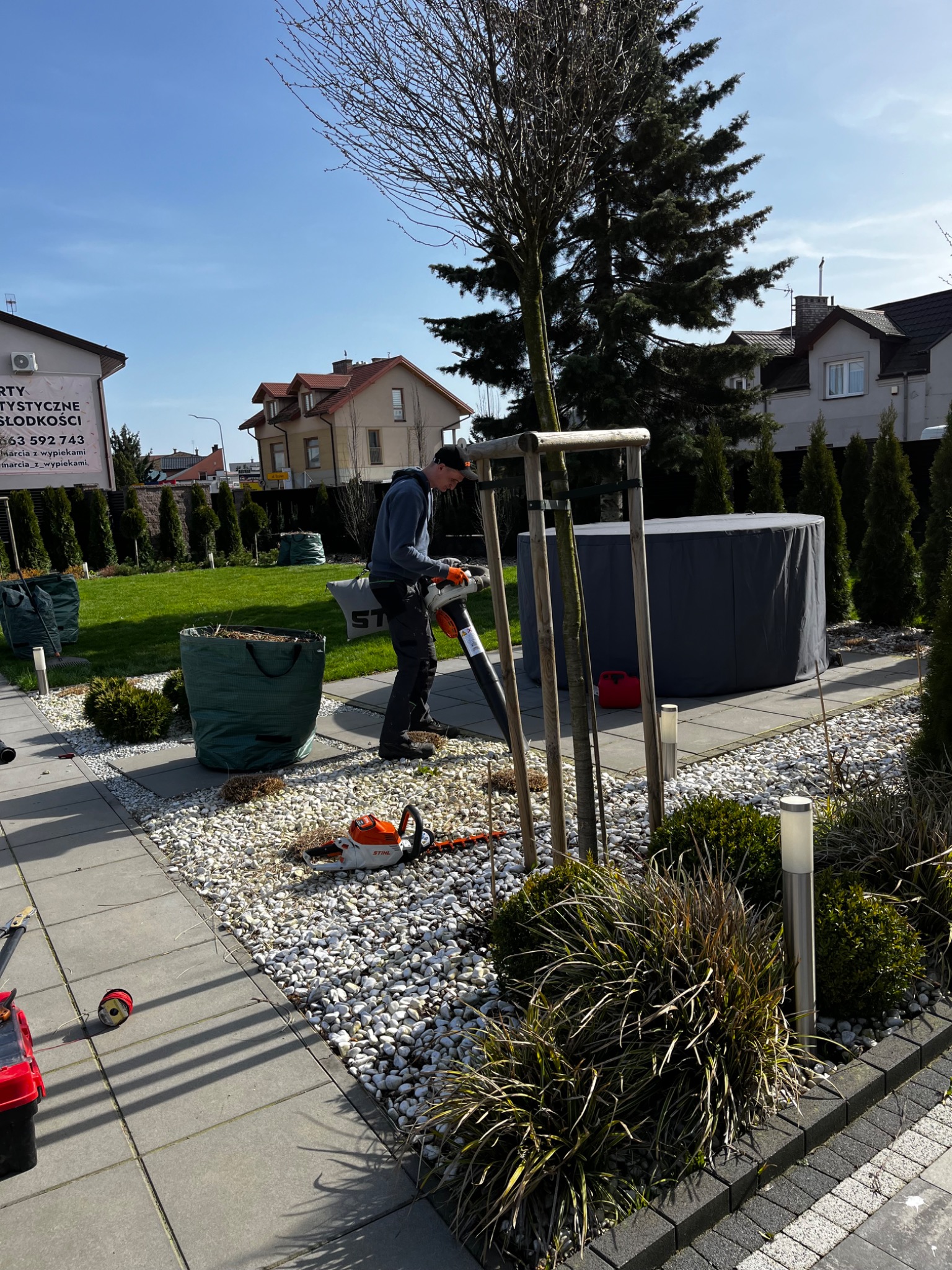 Pracownik w ogrodzie używa dmuchawy do liści, obok leży pomarańczowa pilarka spalinowa Stihl, w tle widoczne worki z odpadami ogrodowymi i zadbany trawnik.